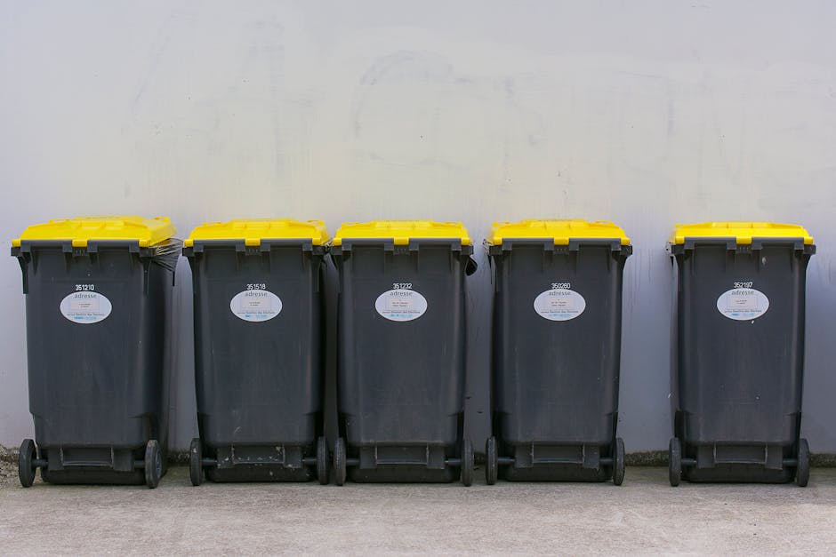 A row of large, upright commercial rubbish bins made of hard plastic, lined up along a pavement beside a brick wall, with each bin featuring a distinct color lid including red, blue, and green. The gr